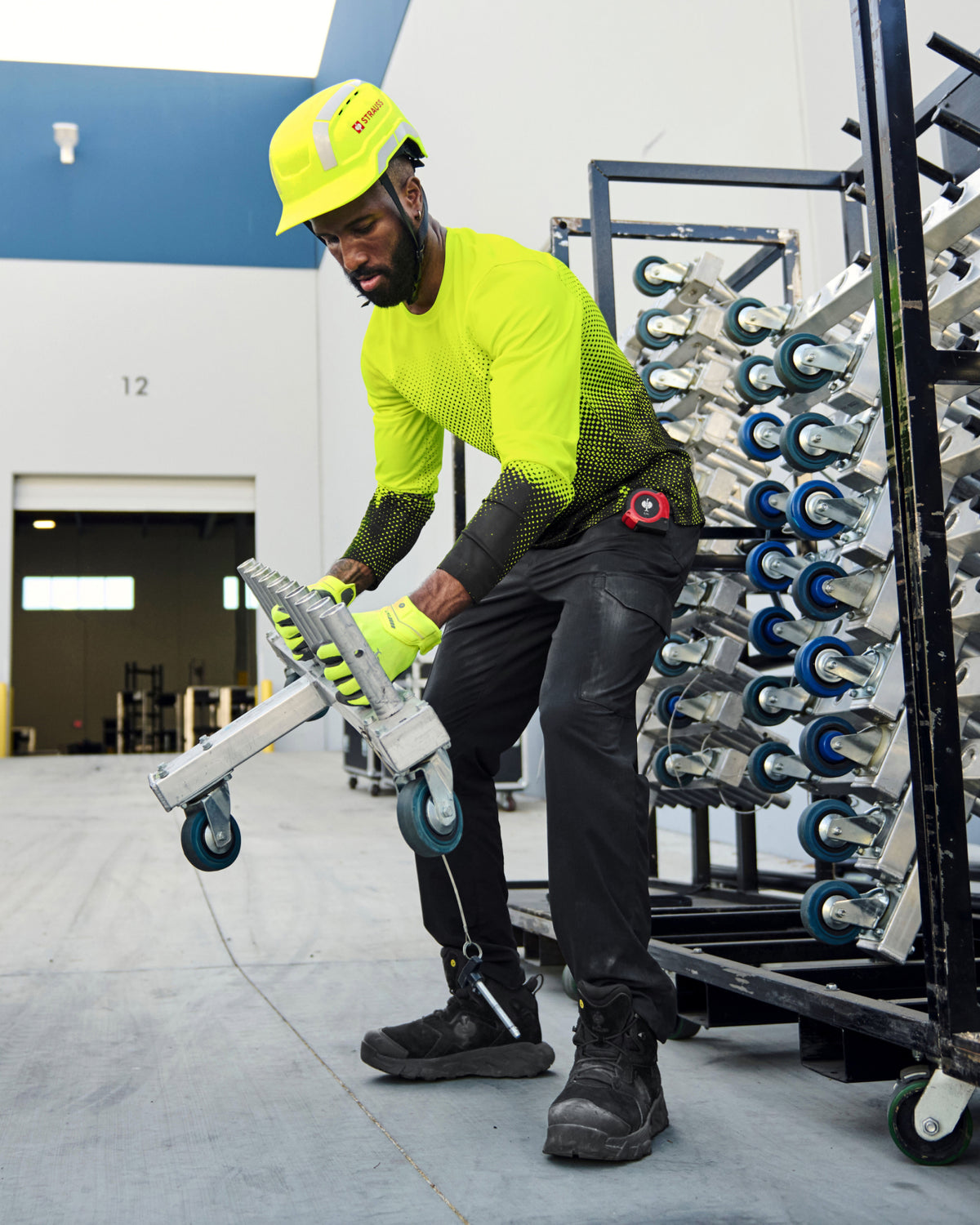 A worker wears the STRAUSS Enhanced Visibility Performance Long Sleeve Shirt e.s.t:aktik in neon yellow and black with black work pants near equipment on a paved jobsite