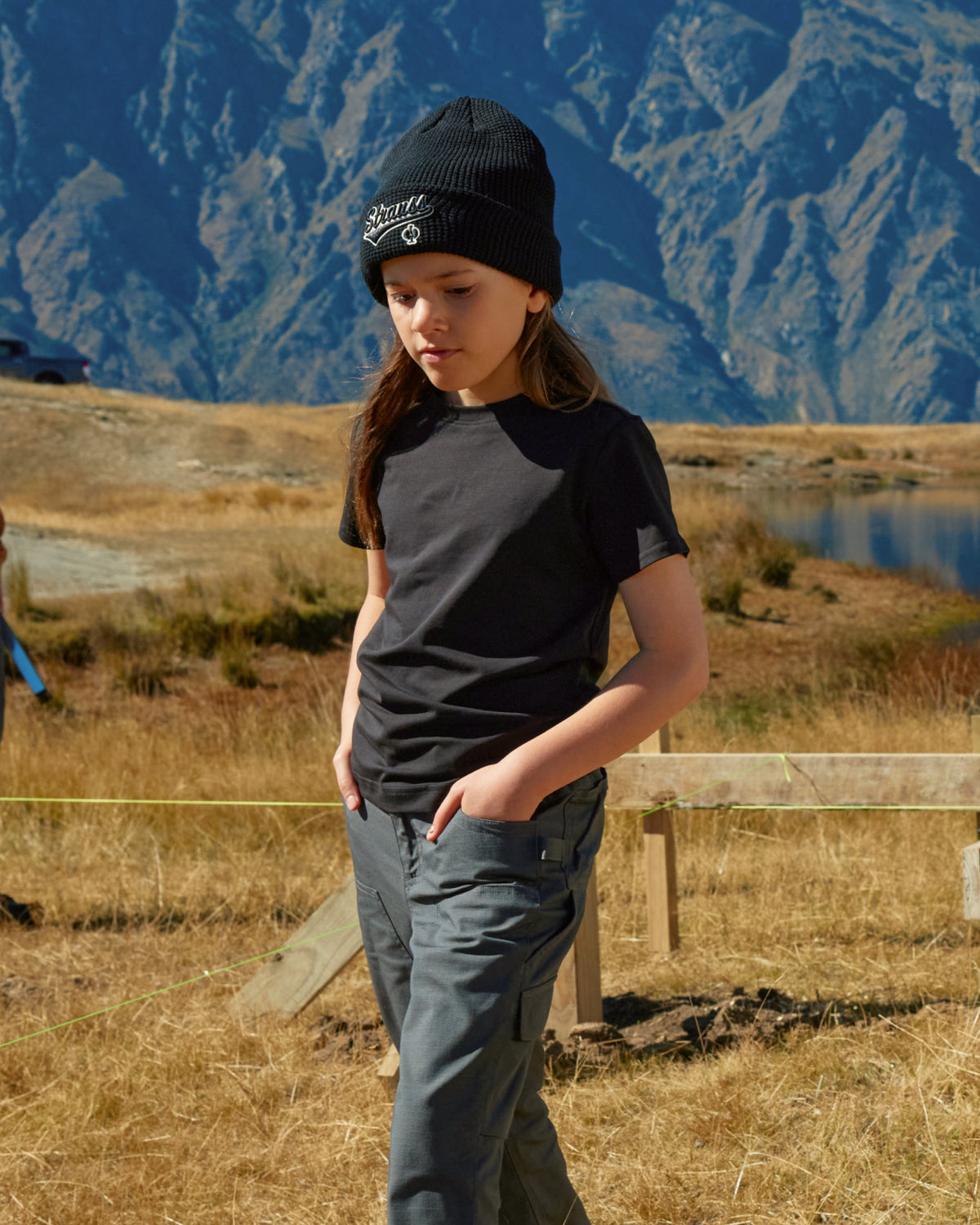 A child wears the Kids' STRAUSS Cotton Stretch T-Shirt in black with kids' work pants beside outdoor work gear