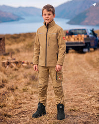 A young boy stands outdoors in a dry grassy field with mountains and a lake in the background, wearing a tan zip-front jacket and matching cargo pants with black boots. A pickup truck is blurred in the background.