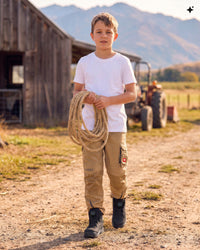 A young boy stands on a dirt path at a farm holding a bundle of rope. He is wearing a white t-shirt, tan cargo pants, and black boots, with a wooden barn, tractor, and mountains in the background.