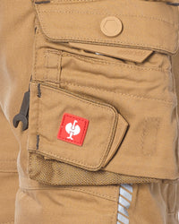Close-up of tan canvas cargo pants showing a front utility pocket with a red STRAUSS patch featuring a white rooster logo, a snap-tab flap, and a small black hook tool tucked into the pocket.