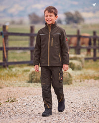 A young boy smiling while walking outdoors on a gravel path, wearing a dark olive brown technical outdoor jacket and matching cargo pants with tan zipper accents.