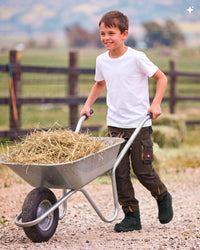 A young boy smiling while pushing a metal wheelbarrow filled with straw on a gravel path in an outdoor farm setting.