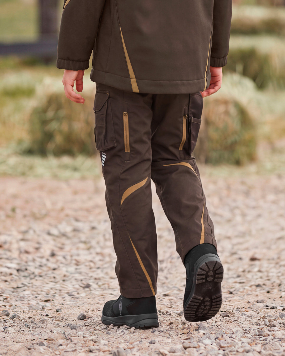 A person outdoors wearing dark brown technical cargo pants with tan contrast stitching and zippered pockets, along with black boots, shown from the back as they walk on a gravel path.