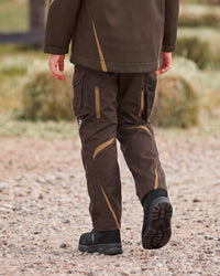 A person outdoors wearing dark brown technical cargo pants with tan contrast stitching and zippered pockets, along with black boots, shown from the back as they walk on a gravel path.
