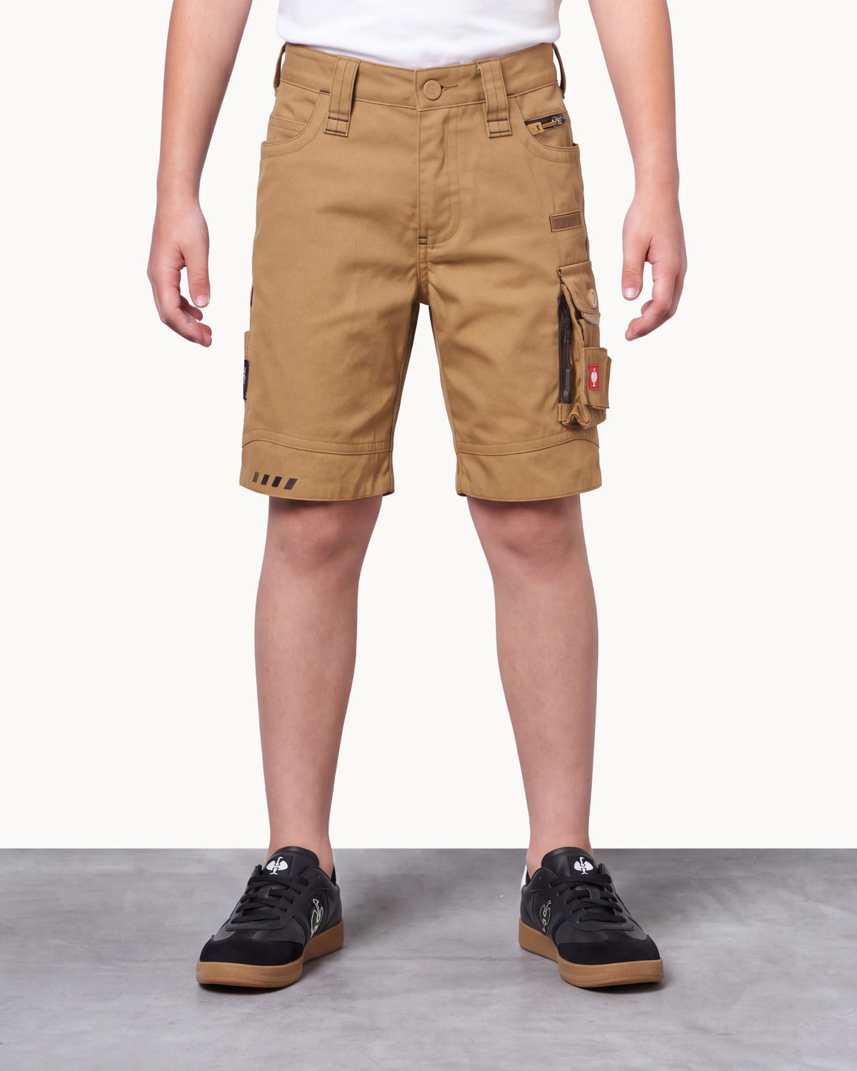 A person wearing tan cargo shorts with rolled cuffs, side cargo pockets, and black low-top sneakers, shown from the waist down against a light studio background.