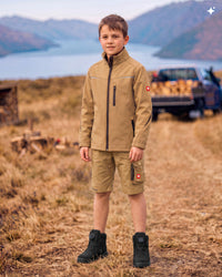 A young boy stands outdoors in a dry grassy field with mountains and water in the background, wearing a tan waterproof jacket, matching shorts, and black boots. A pickup truck is visible in the blurred background.