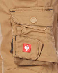 Close-up of a tan canvas utility shorts with a flap pocket featuring a tan snap button and a lower cargo pocket with a red square patch containing a white stylized logo.