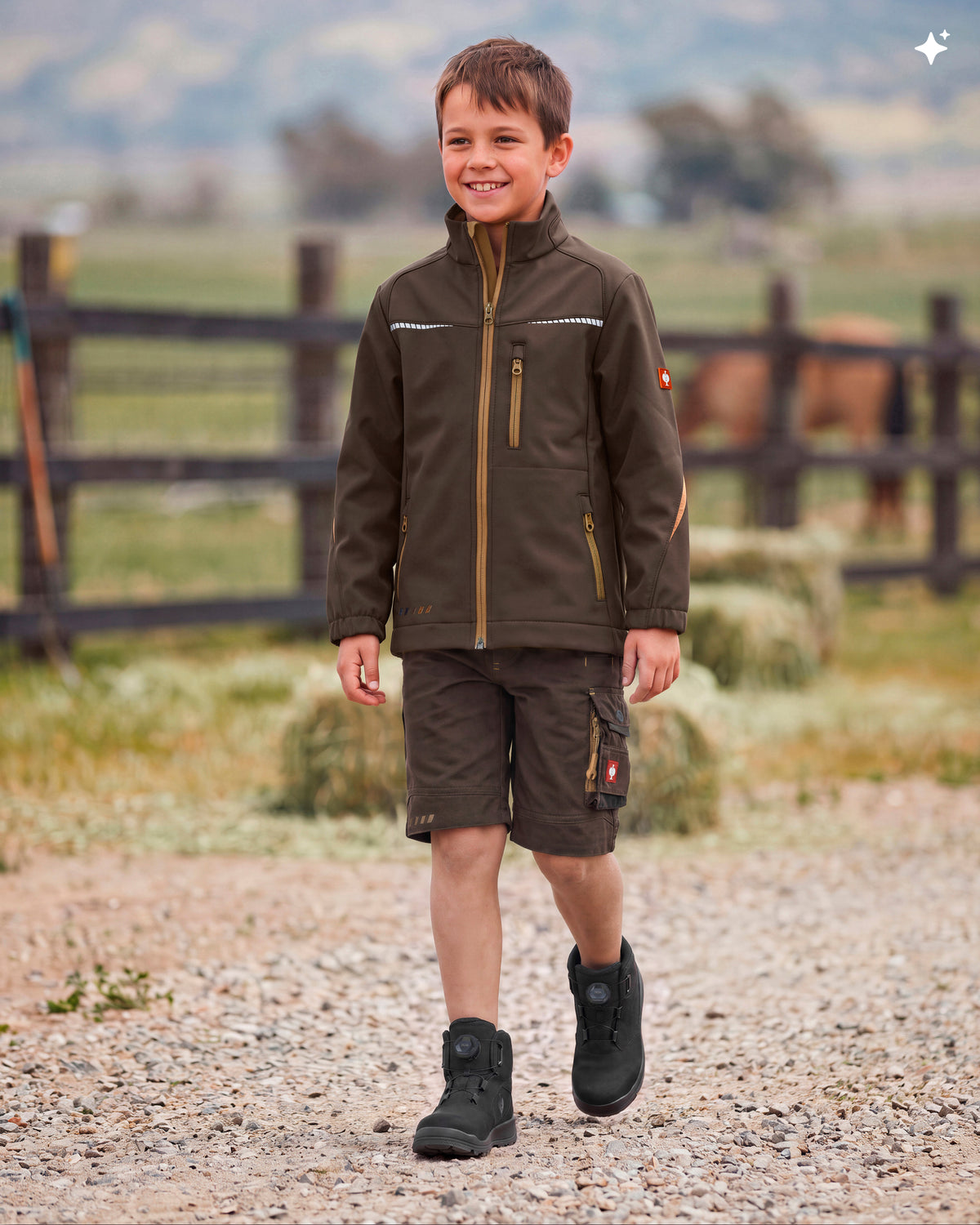 A young boy smiling while walking outdoors on a gravel path, wearing a dark brown technical outdoor jacket with gold-toned zippers and cargo shorts, set against a blurred rural background with a fence and horse.