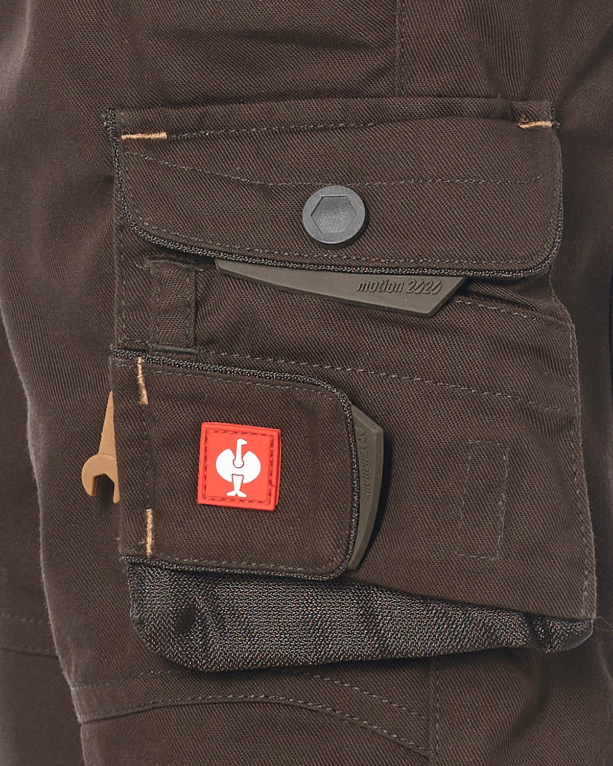 Close-up of a dark brown shorts cargo pocket with a red square patch featuring a white rooster logo, a gray snap button, and a gray metal tool clip.
