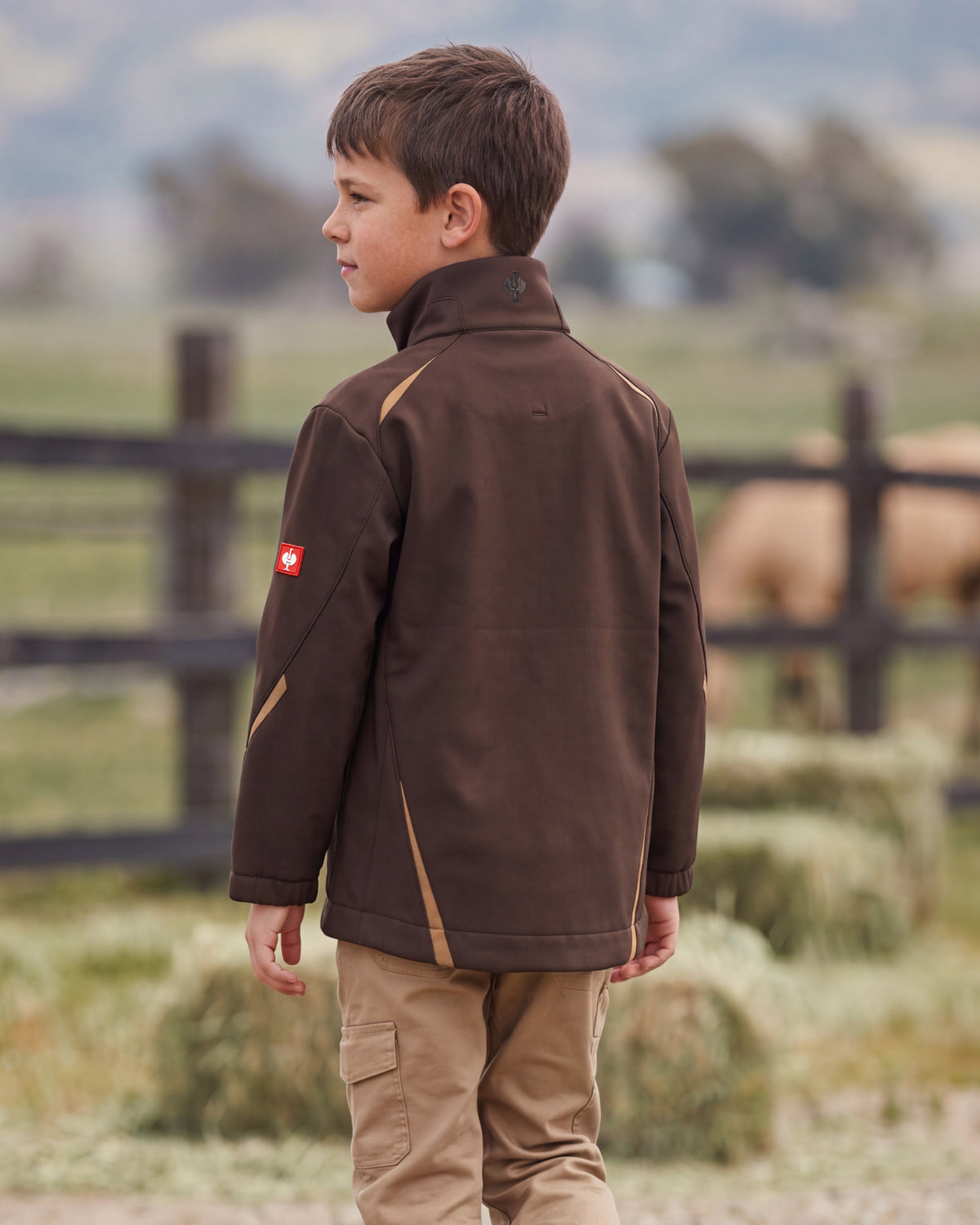 A young boy standing outdoors in a rural setting, wearing a dark brown technical jacket with tan contrast panels and a small red Swiss flag patch on the sleeve, paired with tan pants.