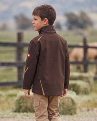 A young boy standing outdoors in a rural setting, wearing a dark brown technical jacket with tan contrast panels and a small red Swiss flag patch on the sleeve, paired with tan pants.