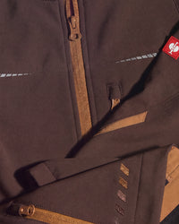 Close-up of a dark brown technical jacket with tan zipper details, a small red logo patch, and reflective striping.