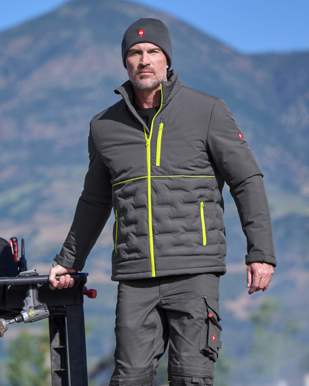 A man wearing a dark gray puffer jacket with bright yellow-green zipper accents, a matching beanie, and dark cargo pants, standing outdoors with a mountain range in the background.