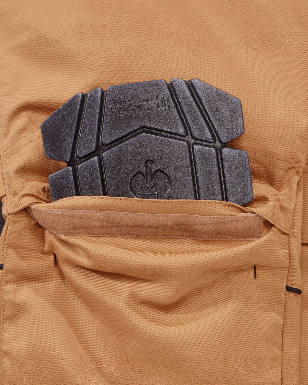 Close-up of a tan technical pants pocket with a black protective knee pad inserted, showing fabric texture and branding embossed on the pad.