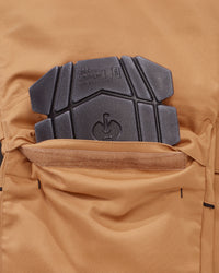 Close-up of a tan technical pants pocket with a black protective knee pad inserted, showing fabric texture and branding embossed on the pad.