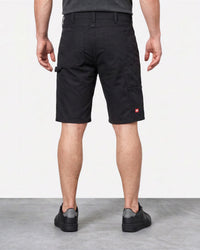 A man viewed from the back wearing black cargo shorts, a dark gray t-shirt, and black sneakers, standing against a white wall and gray floor.