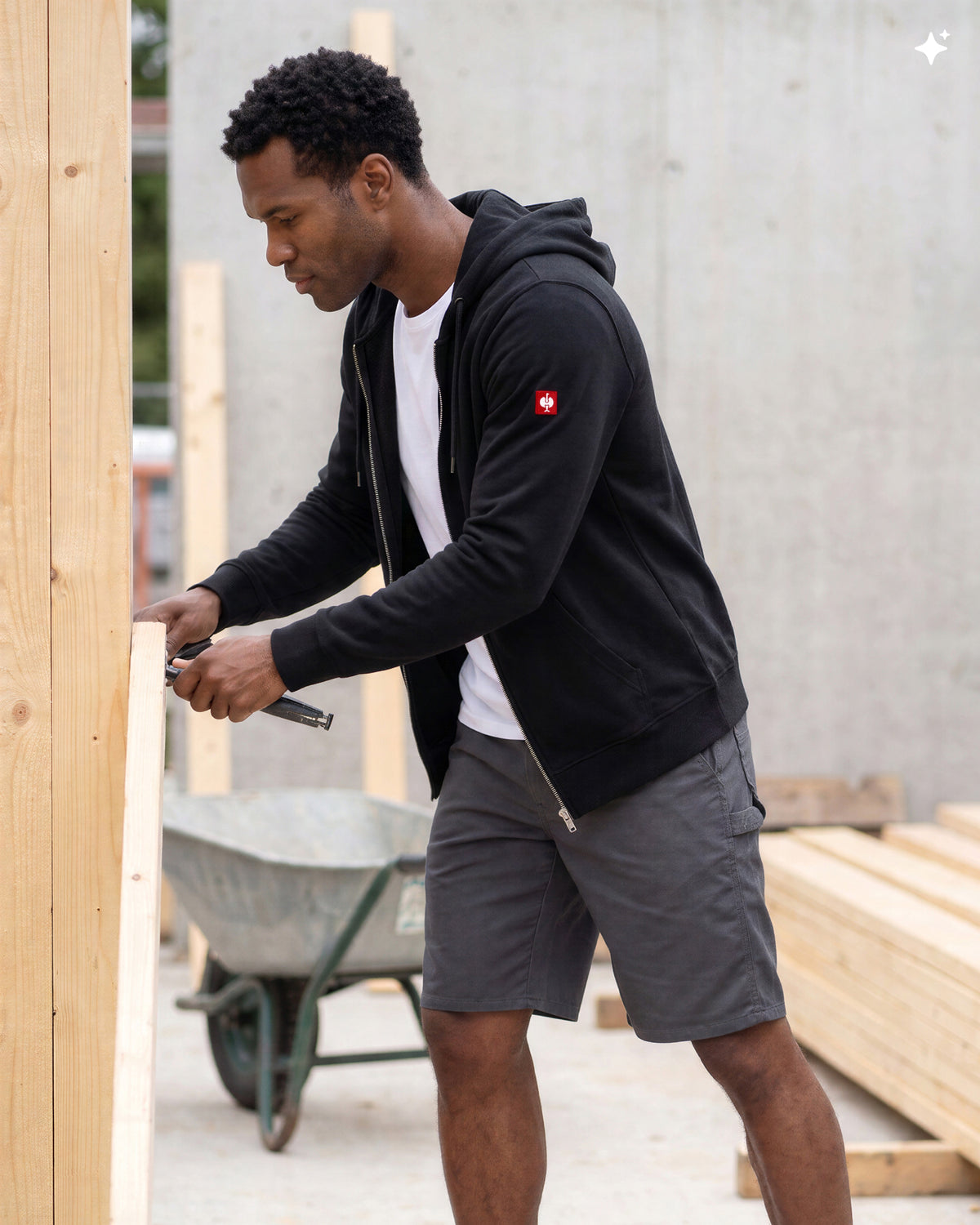 A man wearing a black zip-up hoodie with a small red logo on the sleeve, a white t-shirt, and gray shorts, working on a wooden structure in an outdoor construction setting.