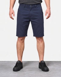 A person wearing navy blue knee-length shorts, a dark gray t-shirt, and black sneakers, standing against a white wall and gray floor.