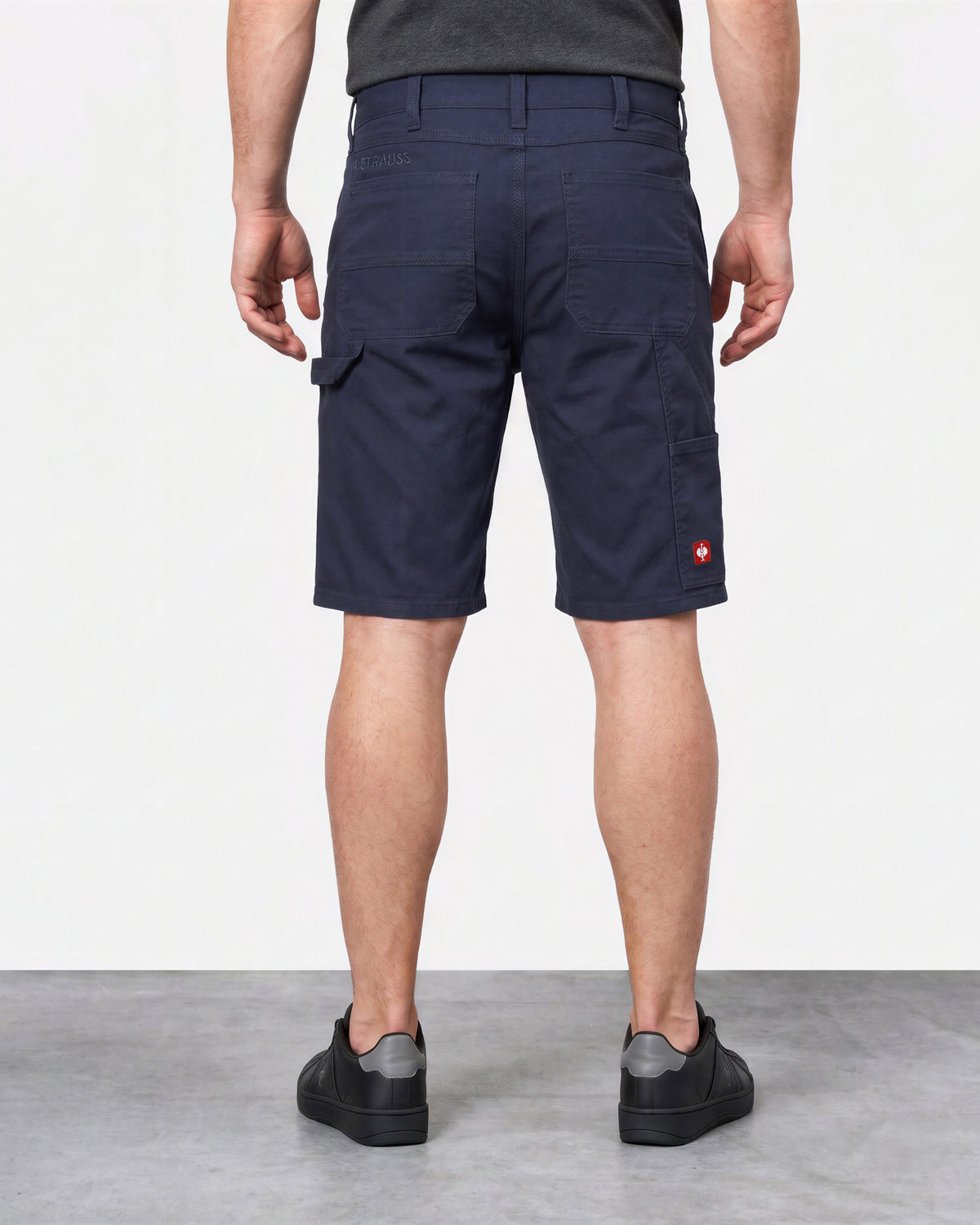 A man viewed from the back wearing navy blue cargo shorts, a dark gray t-shirt, and black sneakers, standing against a light gray studio background.