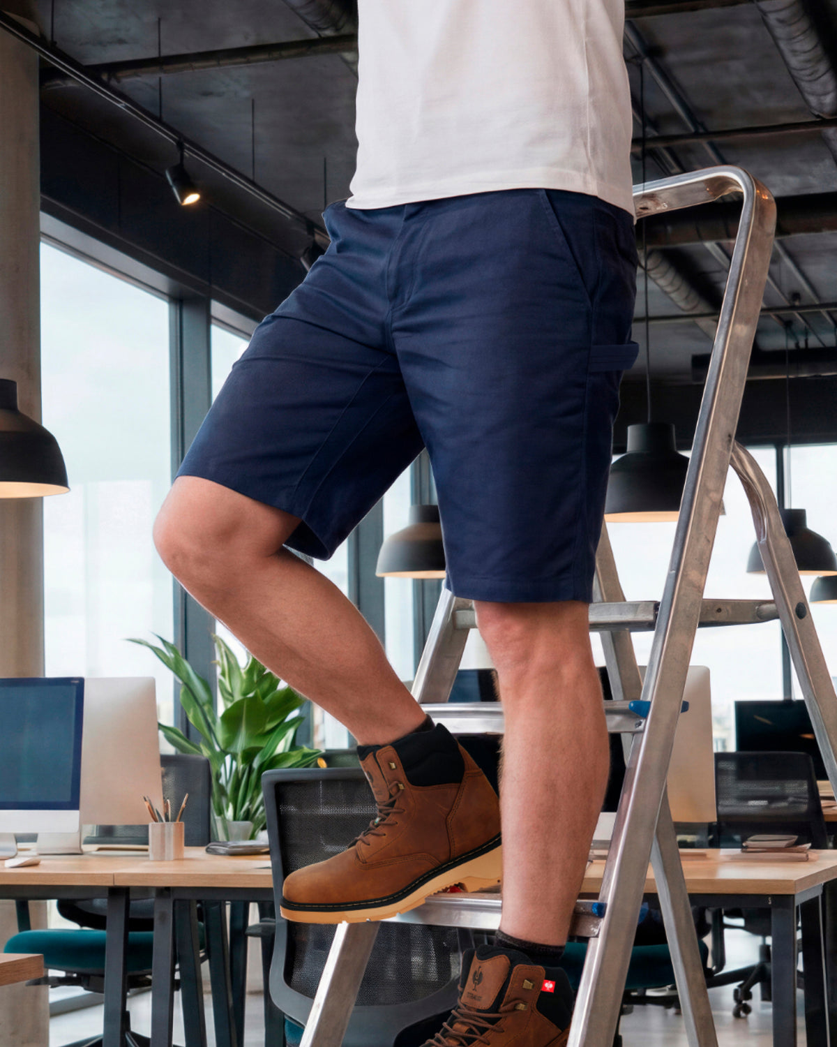 A person wearing navy blue shorts and brown work boots stands on a metal step ladder inside a modern office space with desks, computers, and large windows in the background.