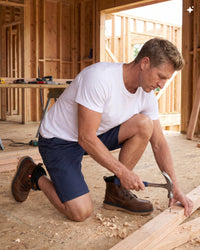 A man kneeling on a wood floor inside a house under construction, using a hammer to secure a piece of wood. He is wearing a white t-shirt, navy blue shorts, and brown work boots.