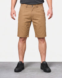 A person wearing tan chino-style shorts, shown from the waist down, standing against a light gray studio background with a gray floor. The shorts are the product being featured, paired with a dark gray shirt and black sneakers.