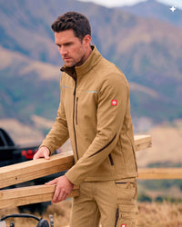 A man wearing a tan technical jacket with a high collar, zippered chest, and a small red logo patch on the upper sleeve, posed outdoors against a blurred mountain background.
