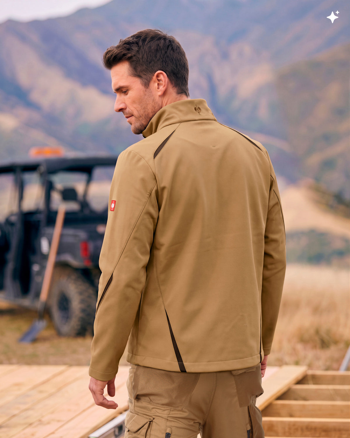 A man wearing a tan technical jacket and matching pants, photographed from the back in an outdoor setting with mountains and a vehicle in the background.