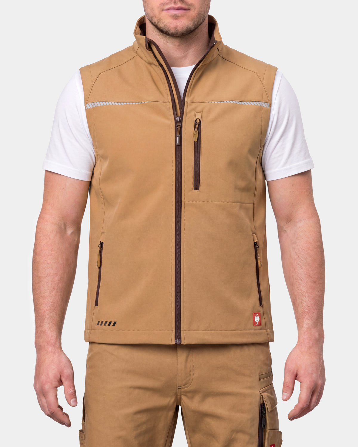 A man wearing a tan zip-up vest over a white t-shirt and matching tan cargo pants, shown from the neck down against a light gray studio background.