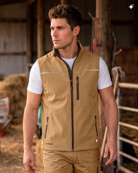 A man wearing a tan zip-front vest over a white t-shirt, standing in a barn setting.