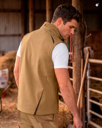 A man wearing a tan insulated vest over a white t-shirt, standing in a rustic barn setting.