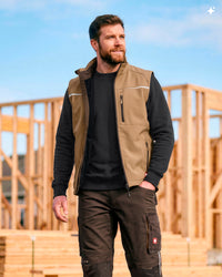 A man wearing a tan and black insulated work vest over a black long-sleeve shirt and brown cargo pants, standing outdoors in front of wooden framing under a blue sky.