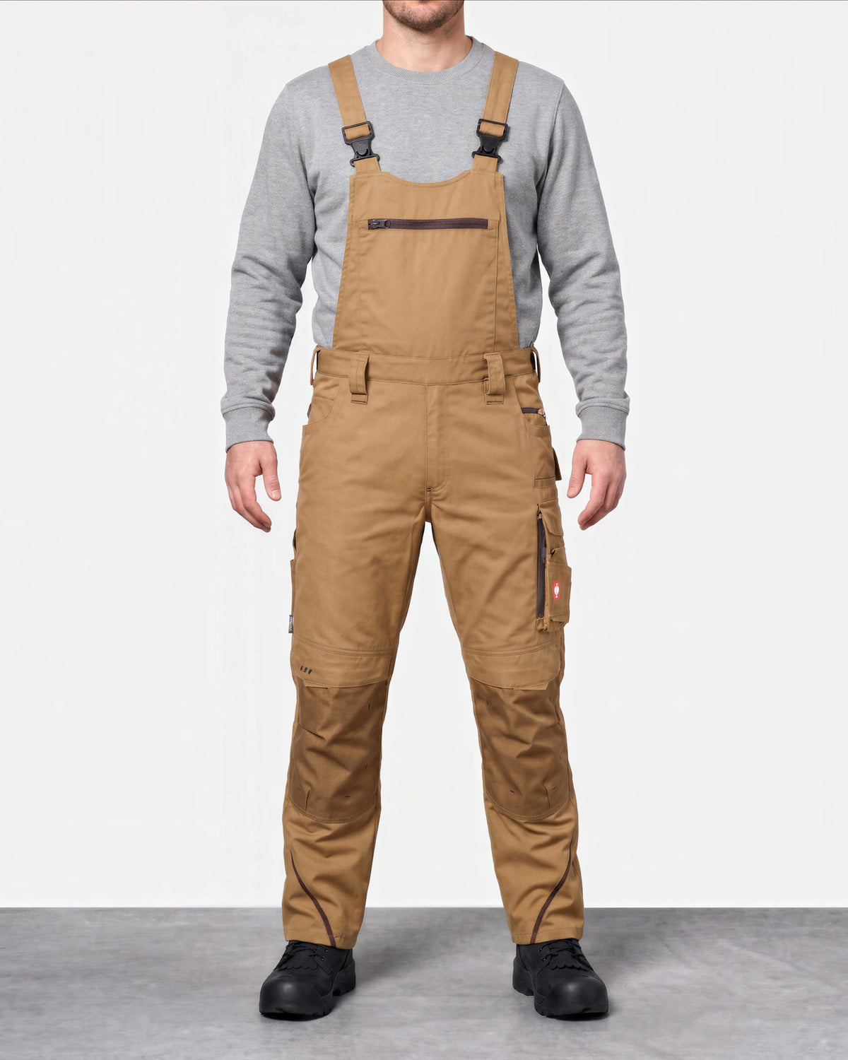 A man wearing tan work bib overalls over a gray long-sleeve shirt, standing against a light gray studio background.