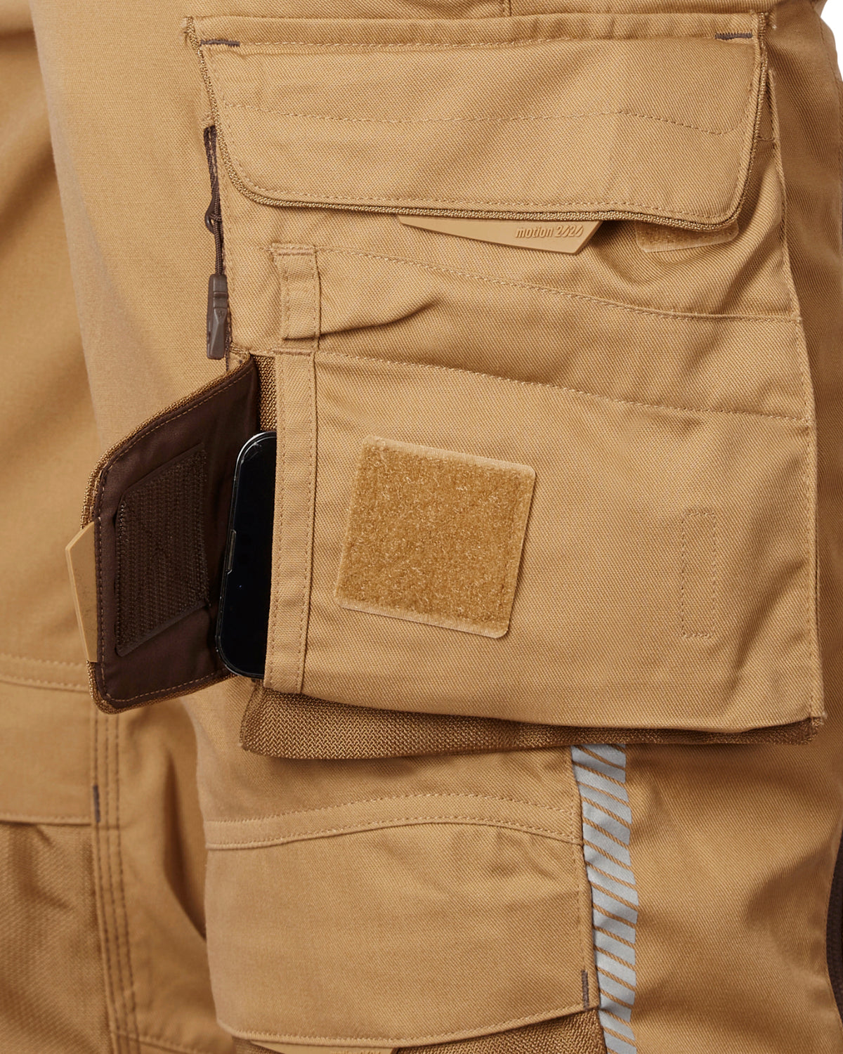 Close-up of a tan cargo bib showing a side utility pocket with a brown zippered card holder containing a black smartphone, alongside a tan Velcro patch.