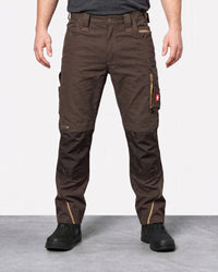 A person wearing dark brown work pants with reinforced knee panels and black work boots, standing against a light gray studio background.