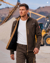A man wearing a dark brown softshell work jacket with tan lining over a white t-shirt, standing in an outdoor construction environment with machinery in the background.