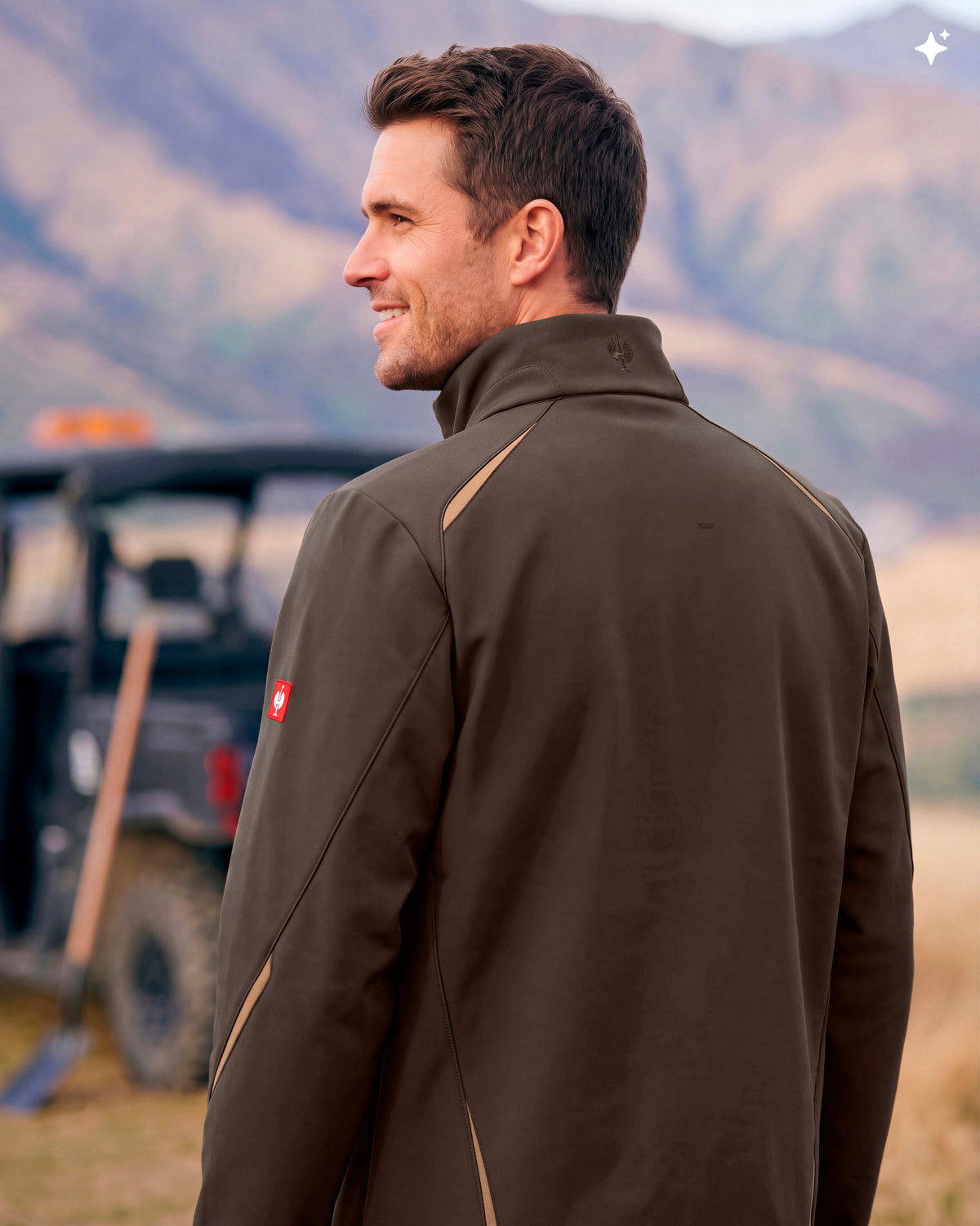 A man wearing a brown athletic jacket with tan contrast stitching, photographed outdoors with a blurred mountain background and a vehicle behind him.