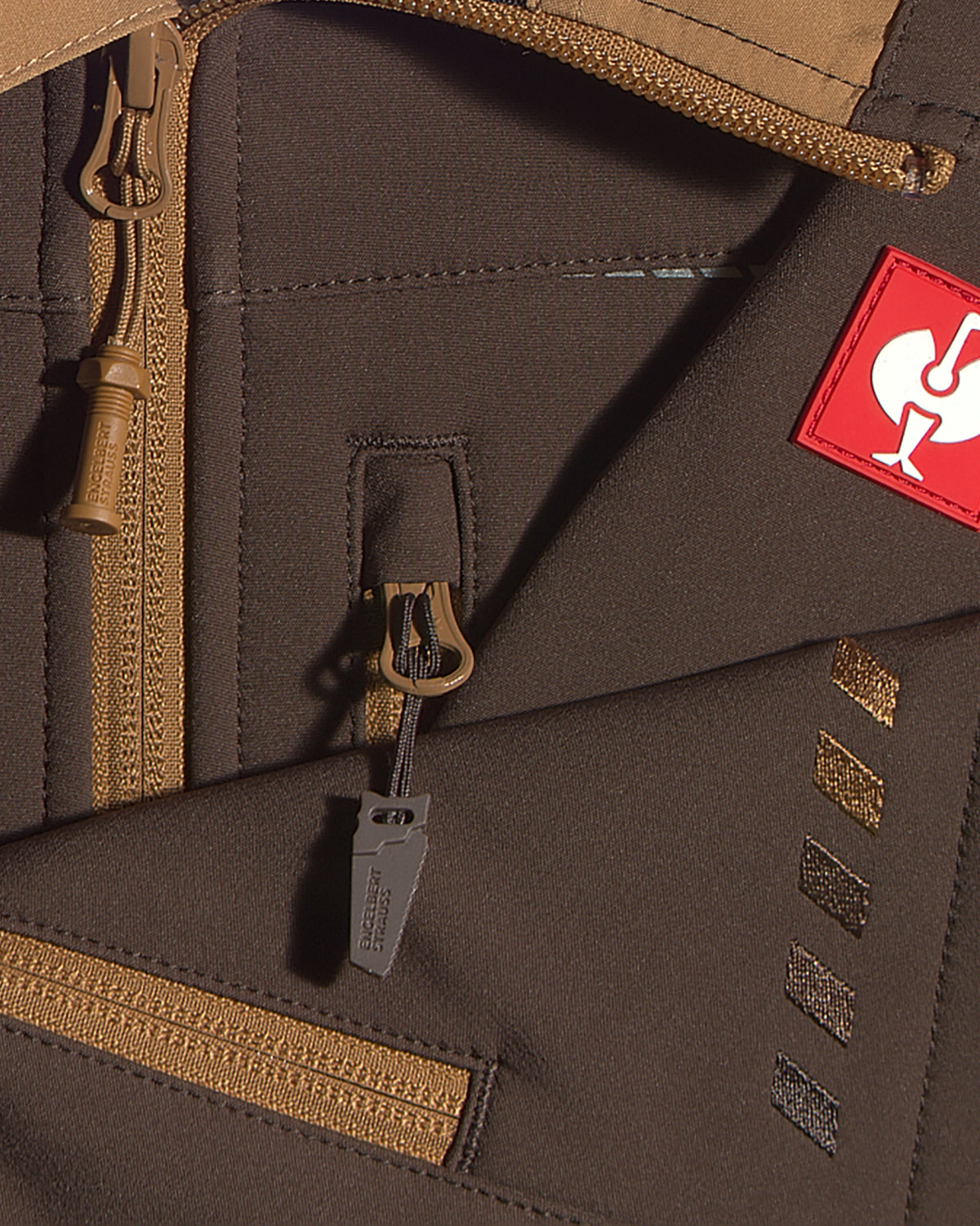 Close-up detail of a brown softshell jacket with tan zippers, a red square patch with a white rooster logo, and a gray zipper pull tag.