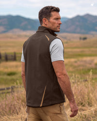 A man wearing a brown sleeveless zip-front vest over a beige short-sleeve shirt and khaki pants, standing in an outdoor field with mountains in the background.