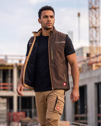 A man wearing a brown work vest over a black t-shirt and khaki cargo pants, standing on a construction site.