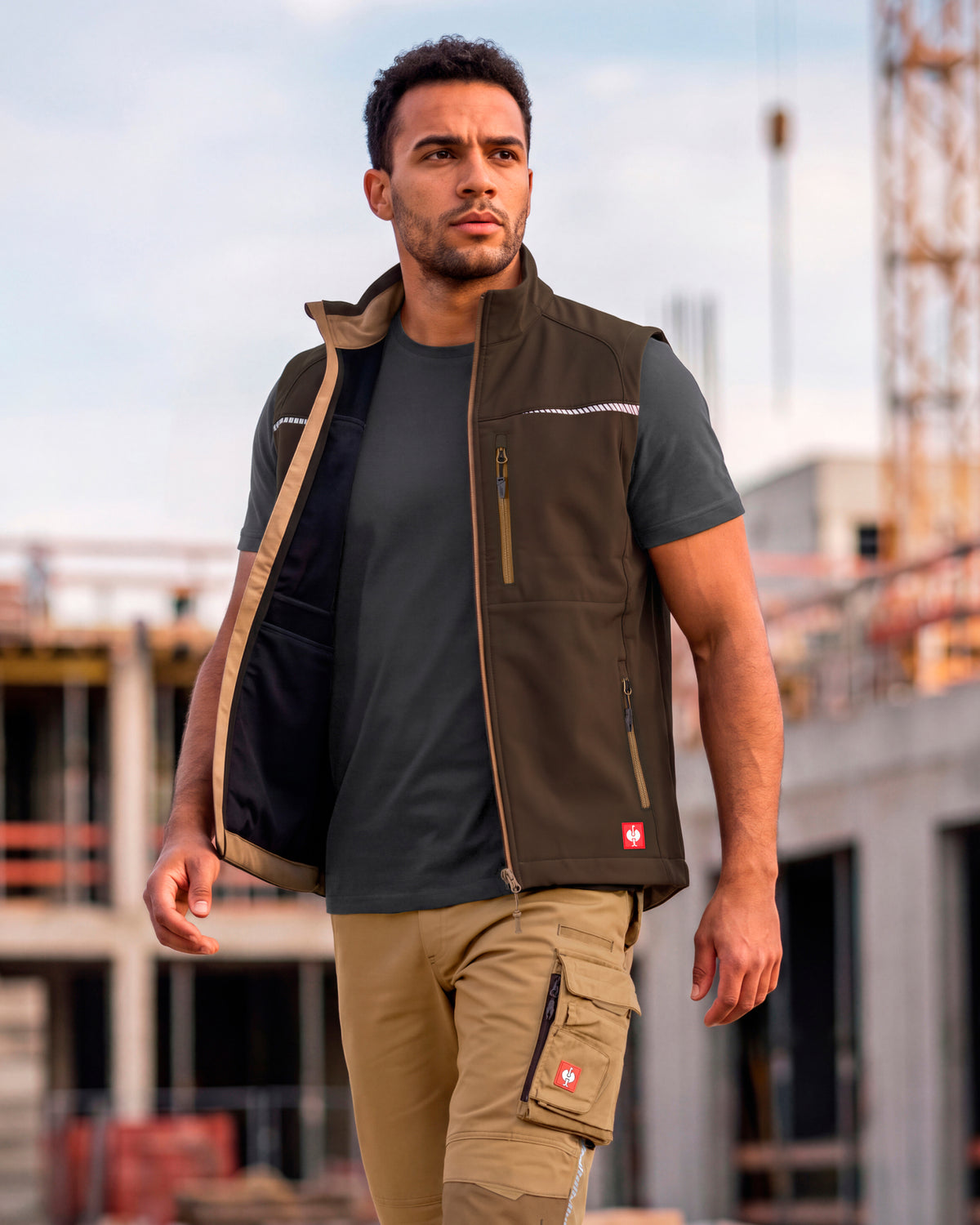 A man wearing a brown work vest over a black t-shirt and khaki cargo pants, standing on a construction site.