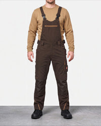 A man wearing dark brown work overalls over a tan long-sleeve shirt and black shoes, standing against a light gray studio background.