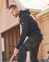 A man in a black hoodie and black work pants bends down holding a shovel in front of a wooden house under construction.