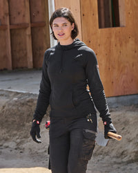 A woman wearing a black hooded work jacket and matching black cargo pants stands outdoors in front of a wooden building, holding a tool in one hand.