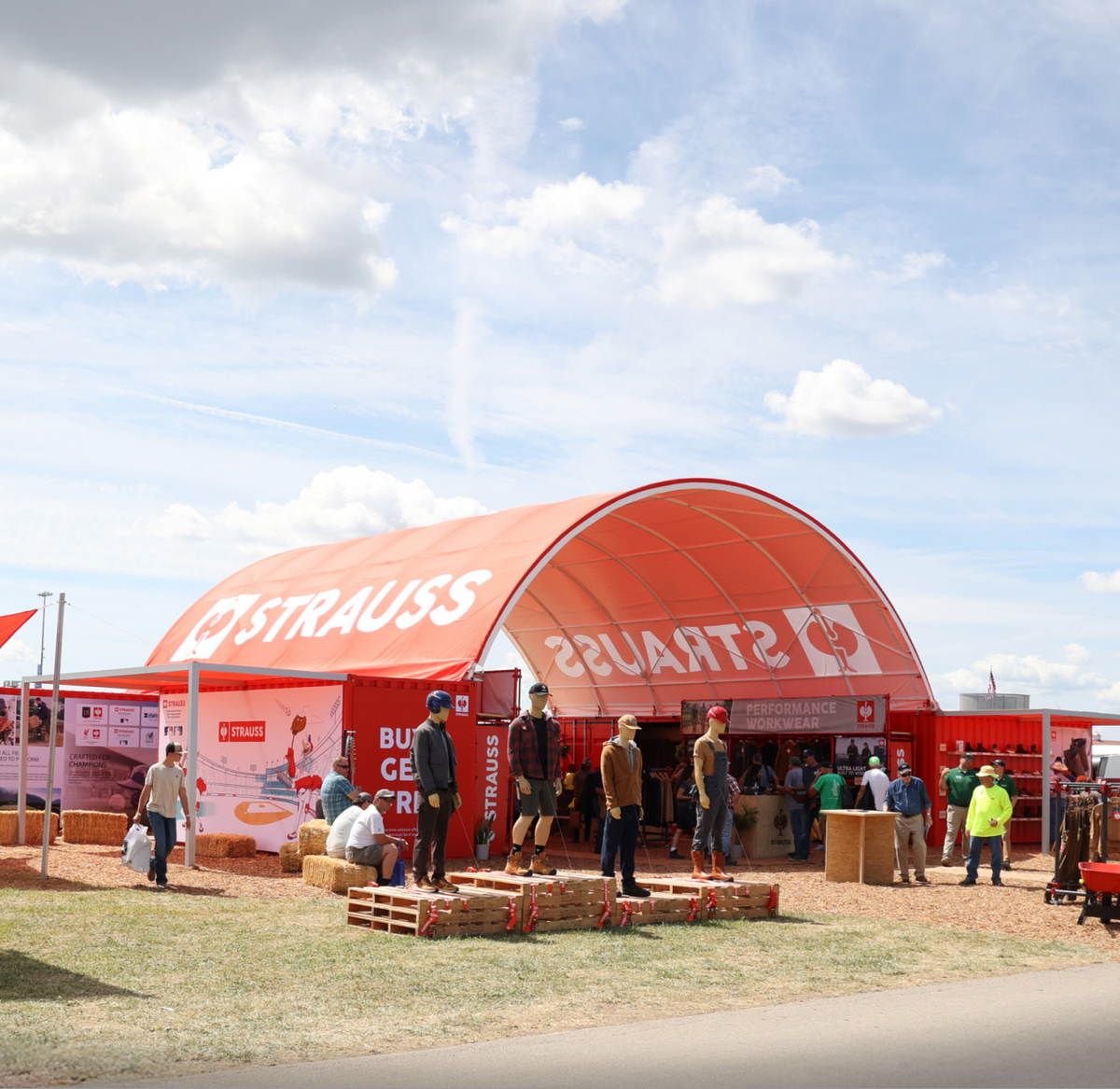 People gather outside a large red tent labeled STRAUSS at an outdoor event. Some are sitting on hay bales, while others stand and walk around under a partly cloudy sky.