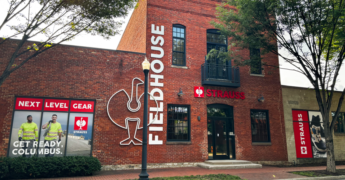 A red brick building labeled Fieldhouse and Strauss with large windows, signage, and two people in safety gear on a poster that says Next Level Gear. Get Ready, Columbus. Trees frame the scene.
