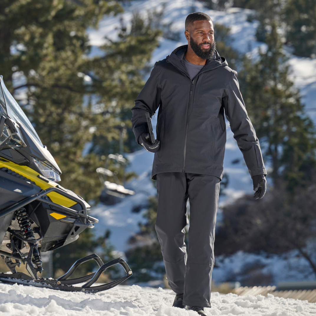 Man in winter clothing standing next to a snowmobile in a snowy landscape with trees.