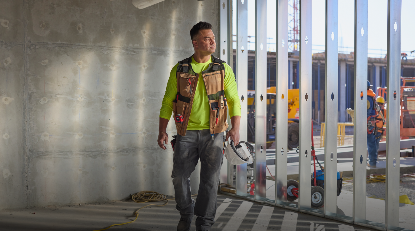 Nick Swisher wearing a bright yellow safety shirt, brown tool vest, and gray work pants walks through a modern interior space with vertical metal panels and a concrete wall.