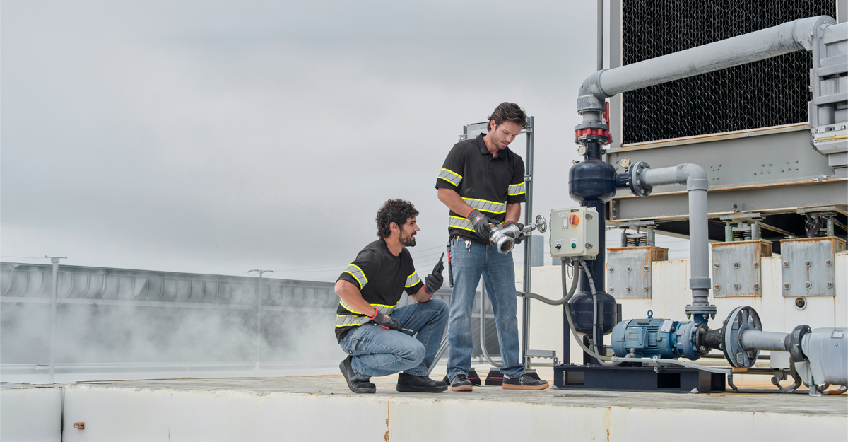 Two workers in reflective shirts inspect and operate industrial equipment on a rooftop, with pipes and machinery visible. Steam rises near them under a cloudy sky.
